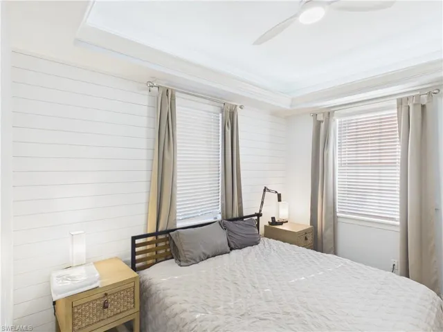 Bedroom featuring shiplap accent wall, tray ceiling with crown molding, ceiling fan, and two windows with blinds