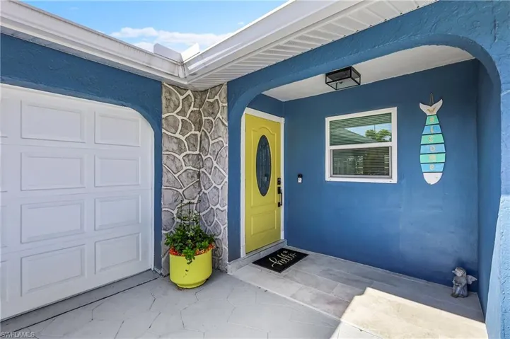 Vibrant blue stucco exterior featuring a sunny yellow front door with an oval glass insert, a panel garage door, a stone veneer accent wall, and a patterned concrete entry pad