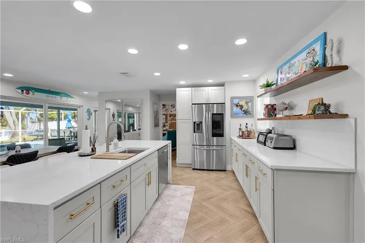 Contemporary kitchen featuring an expansive central island with an undermount sink and modern faucet, white cabinetry with gold-tone hardware, stainless steel appliances, and wood-finish herringbone flooring
