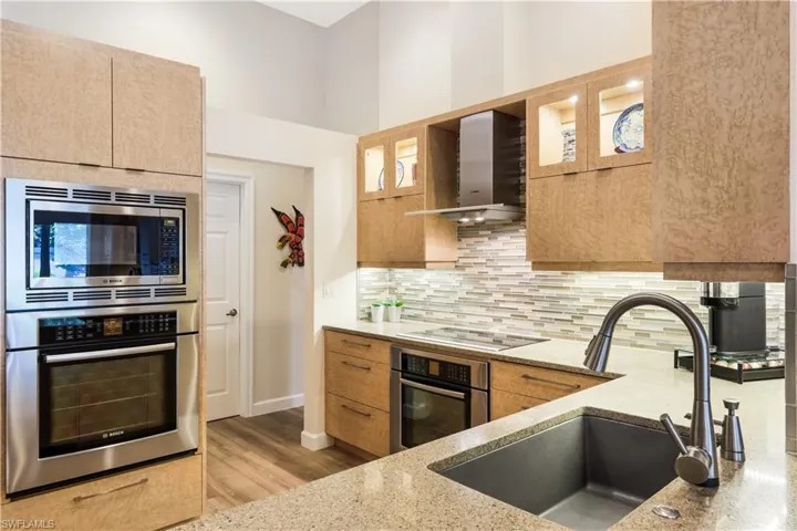 Integrated stainless steel microwave and oven, built-in cooktop, under-cabinet lighting, natural wood-finish cabinetry, and a single-basin sink with a gooseneck faucet