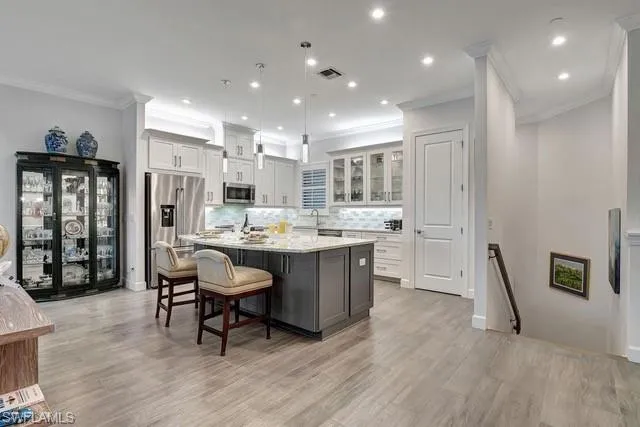 Featuring a large island with seating, hanging pending light fixtures, stainless steel appliances, and cabs with glass fronts and open staircase
