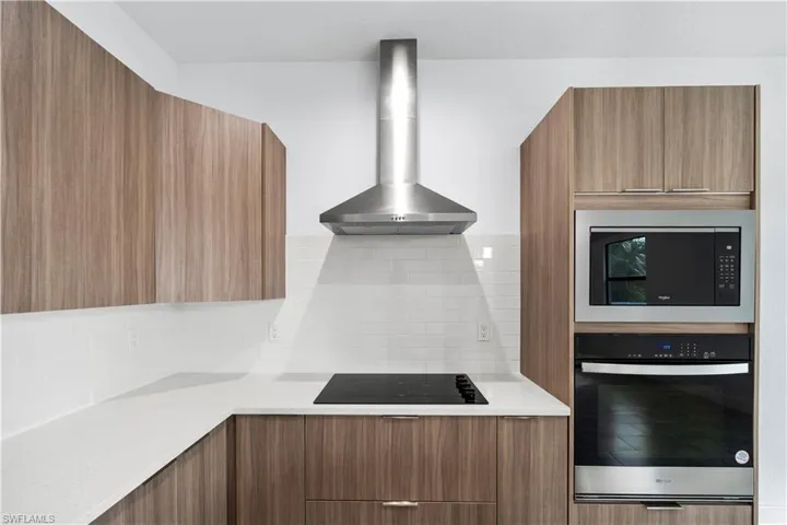 Kitchen with brown cabinetry, modern cabinets, wall chimney exhaust hood, stainless steel appliances, and light stone counters