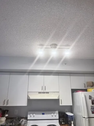Kitchen view of range hood, refrigerator, stove, white cabinetry, and a textured ceiling