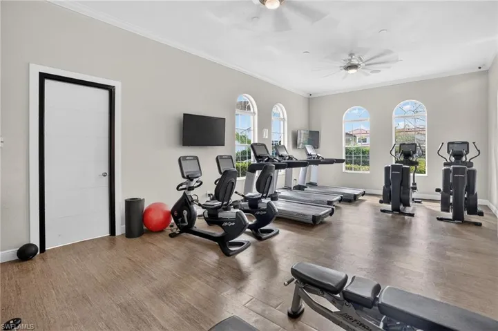 Fitness room featuring wood-finish flooring, three arched windows, two ceiling fans, and two wall-mounted televisions