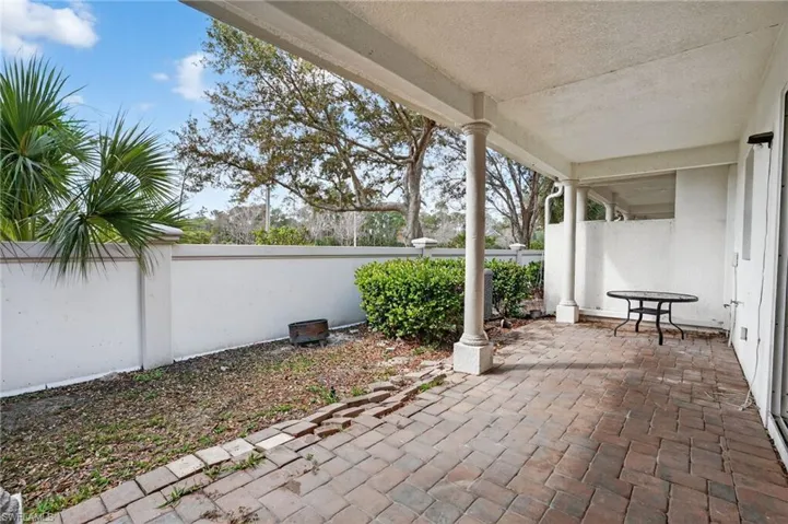 Covered patio featuring brick paver flooring, support columns, a white privacy wall, and mature landscaping