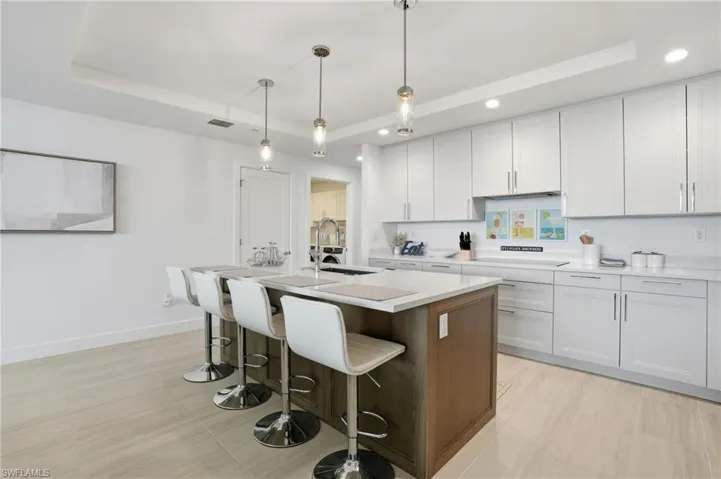 Kitchen featuring a raised ceiling, an island with sink, a kitchen breakfast bar, white cabinetry, and decorative light fixtures - Virtually Edited Image