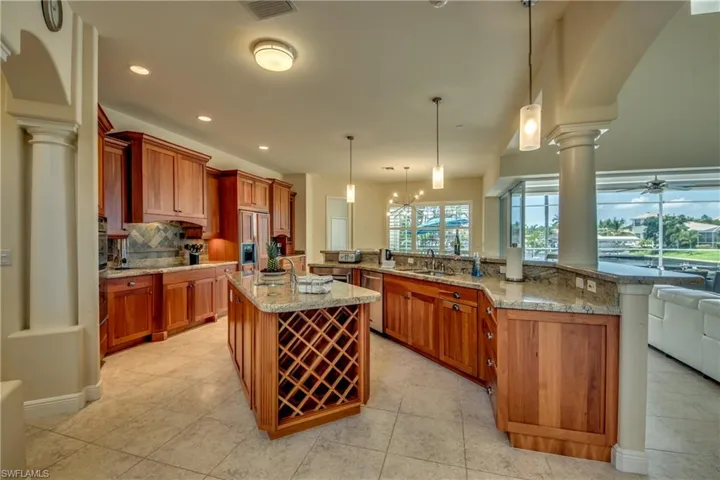 Kitchen with decorative columns, hanging light fixtures, light stone counters, brown cabinetry, and recessed lighting
