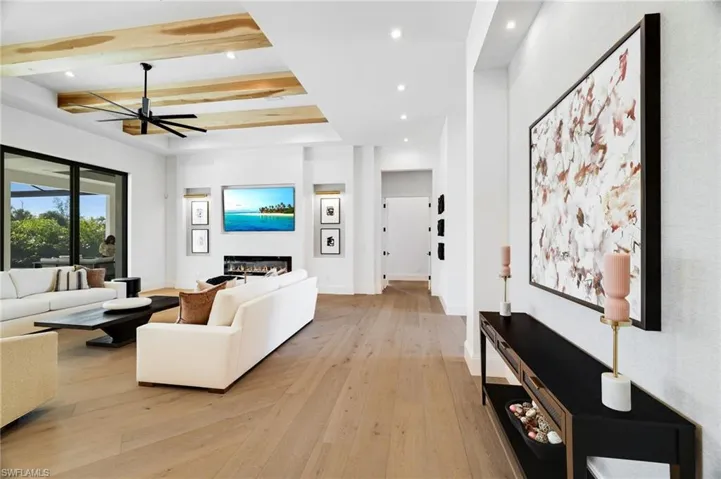 Living area featuring light wood-style floors, a glass covered fireplace, a ceiling fan, recessed lighting, and beam ceiling