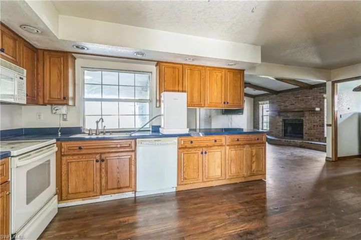 Kitchen with white appliances, sink, a brick fireplace, dark wood-type flooring, and beam ceiling