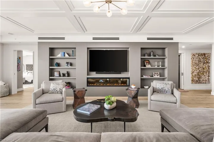 Living room featuring light wood-style floors, built in shelves, a glass covered fireplace, a chandelier, and coffered ceiling
