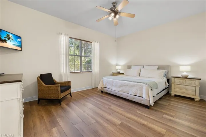 Primary bedroom with woodlike flooring, king bed, side tables, dresser looking onto the rear preserve area.