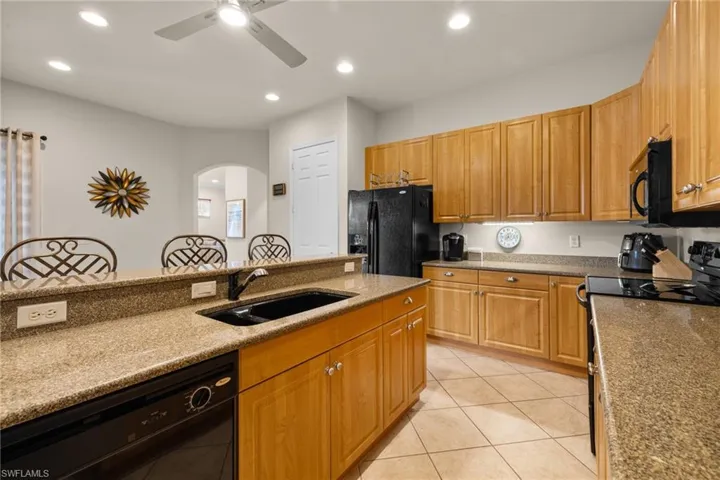 Kitchen island with sink, light tile patterned floors, ceiling fan, black appliances, and light granite countertops