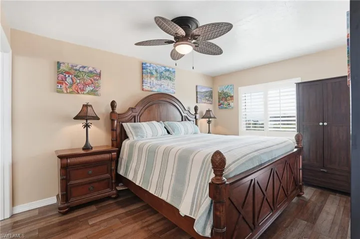 Bedroom featuring wood-finish flooring, a ceiling fan with integrated lighting, and a window with white plantation shutters - Virtually Edited Image