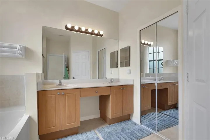 Dual-sink vanity with light wood cabinetry, integrated stone-surface countertop, full-width wall mirror, and mirrored sliding closet doors