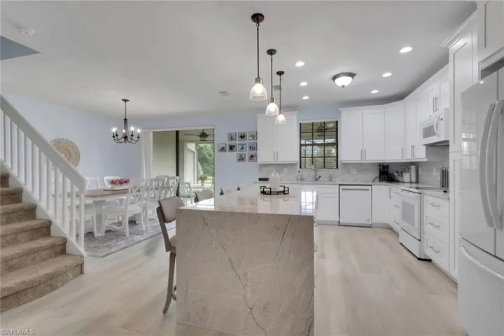 Kitchen featuring white shaker-style cabinetry, an expansive waterfall-edge stone island, wood-finish flooring, and recessed ceiling lighting complemented by three glass pendant lights