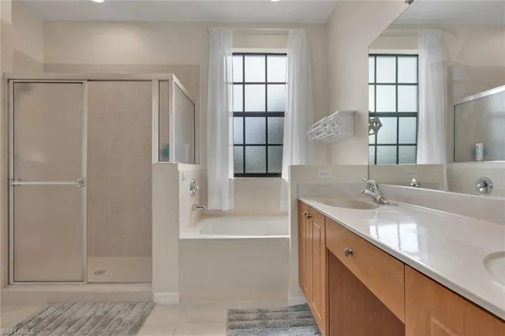 Dual-sink vanity with a light wood finish and solid surface countertop, glass-enclosed shower with tiled surround, and a recessed soaking tub featuring a decorative grid-pane window