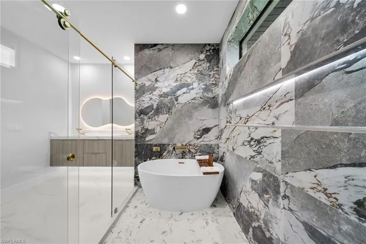 Bathroom featuring double vanity, a freestanding bath, light marble finish flooring, stone wall, and recessed lighting