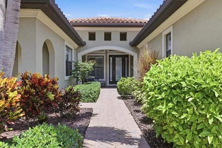 Paver walkway leading to an arched entry with a glass-paneled double door, surrounded by lush landscaping and a tiled roof