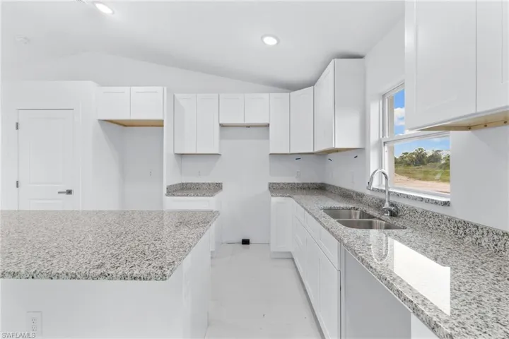 Kitchen with a sink, vaulted ceiling, light stone countertops, white cabinetry, and recessed lighting