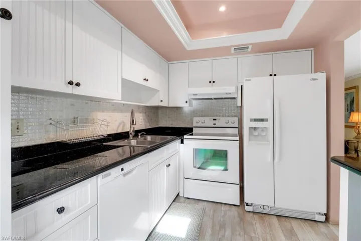 Kitchen with white appliances, under cabinet range hood, a tray ceiling, dark countertops, and decorative backsplash