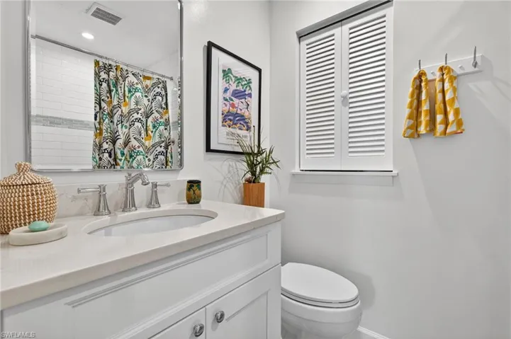 Bathroom featuring a white vanity with a light-toned countertop, a rectangular mirror, a toilet, and a window with white louvered shutters
