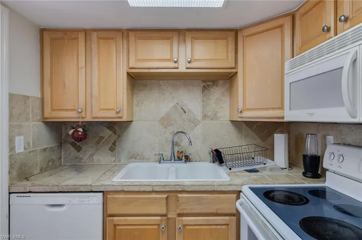 Kitchen featuring white appliances, light wood finish cabinetry, decorative backsplash, and tile countertops