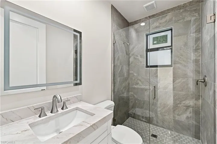 Backlit vanity mirror, undermount sink, stone countertop, glass-enclosed shower, and tiled shower walls