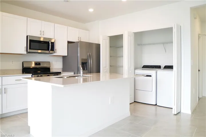 Kitchen featuring stainless steel appliances, white cabinetry, light stone countertops, a kitchen island with sink, and washer and dryer