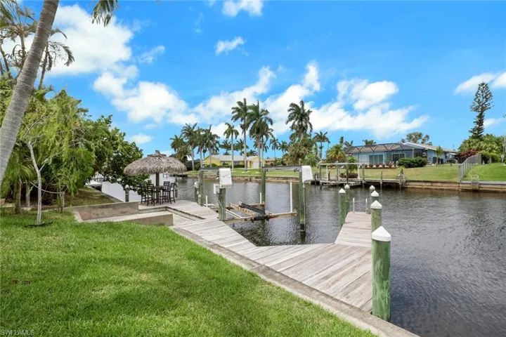 Waterfront property featuring a wooden dock with multiple boat slips, a manicured lawn, tropical landscaping, and a thatched-roof outdoor bar area