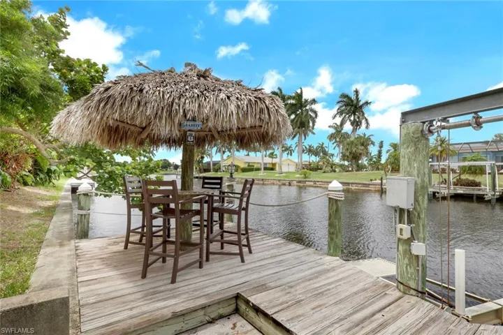 Waterfront dock with a thatched umbrella and seating area, featuring a boat lift and tropical landscaping