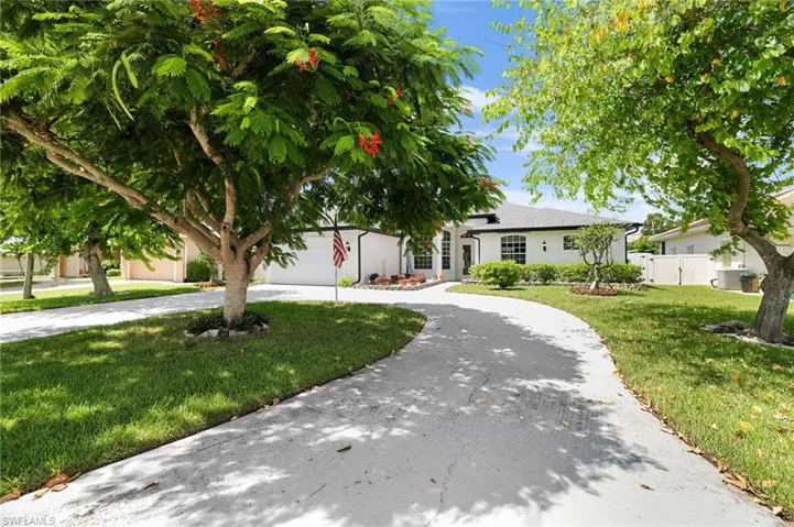 Circular paved driveway, mature trees, landscaped front yard, white exterior finish, and dark roof