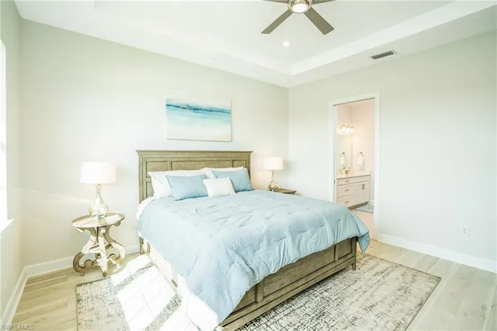 Bedroom featuring light wood-type flooring, a raised ceiling, ensuite bath, a ceiling fan, and recessed lighting