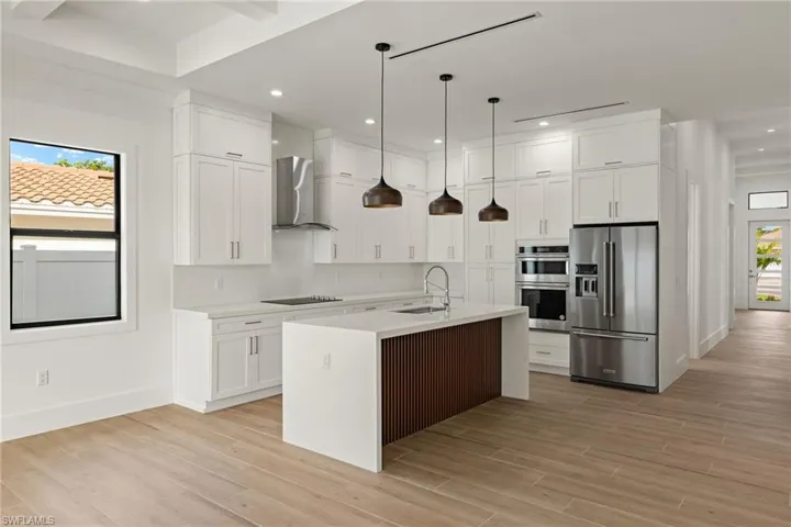 Kitchen with pendant lighting, a kitchen island with sink, wall chimney range hood, appliances with stainless steel finishes, and white cabinetry