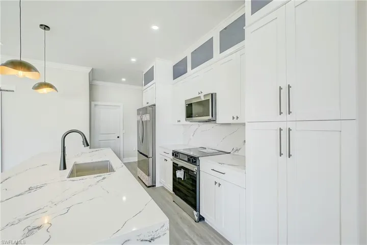 Kitchen featuring hanging light fixtures, appliances with stainless steel finishes, light stone counters, glass insert cabinets, and ornamental molding