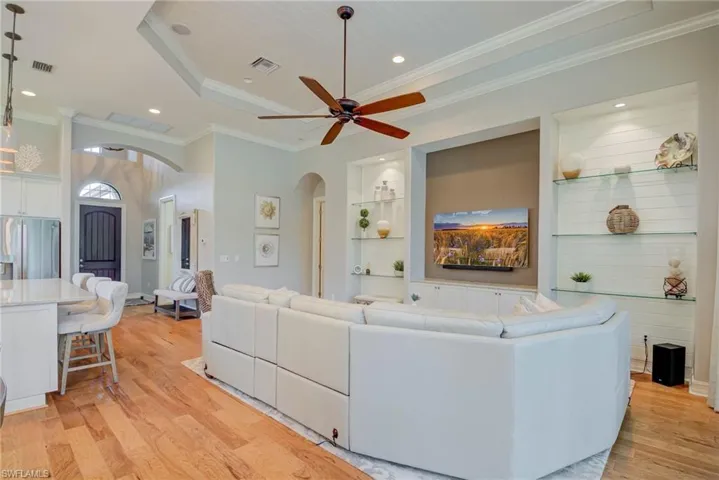 Living room with arched walkways, light wood-type flooring, ceiling fan, crown molding, and a raised ceiling