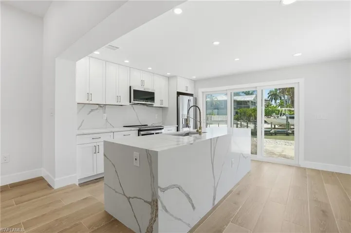 Modern kitchen featuring light wood-style flooring, white cabinetry, a large kitchen island, and a stainless steel refrigerator - Virtually Edited Image