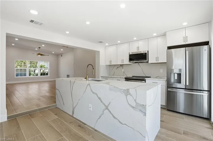 The kitchen features a large island with a waterfall edge and integrated sink, stainless steel appliances, and white cabinetry - Virtually Edited Image