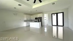 Expansive open-concept living area featuring light-toned tile flooring, a tray ceiling with a ceiling fan, and a contemporary kitchen with white cabinetry and a central island