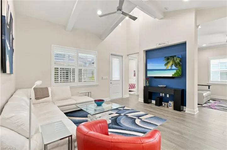 Vaulted ceiling with exposed beams, ceiling fan, window with plantation shutters, wood-finish flooring, and a recessed media wall with accent color