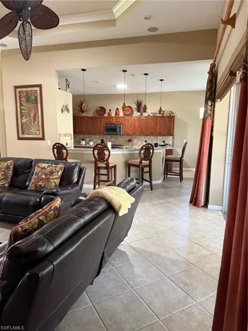 Open-concept kitchen with wood cabinetry, stainless steel microwave, and pendant lighting, overlooking a living area with tile flooring and a decorative ceiling fan