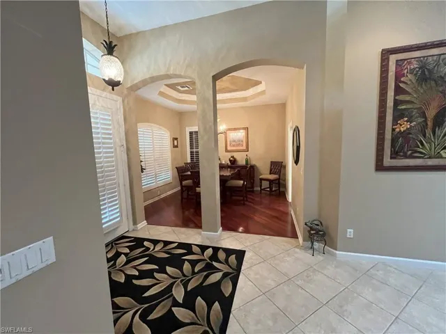 Tiled entryway with an arched doorway opening to an interior space featuring wood-finish flooring, plantation shutters, and a tray ceiling
