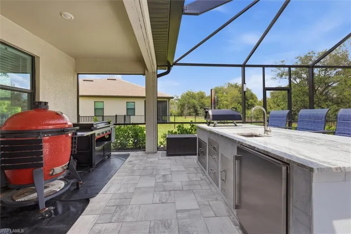 Screened patio featuring a built-in outdoor kitchen with integrated stainless steel appliances, a sink with a gooseneck faucet, and a light-toned stone countertop
