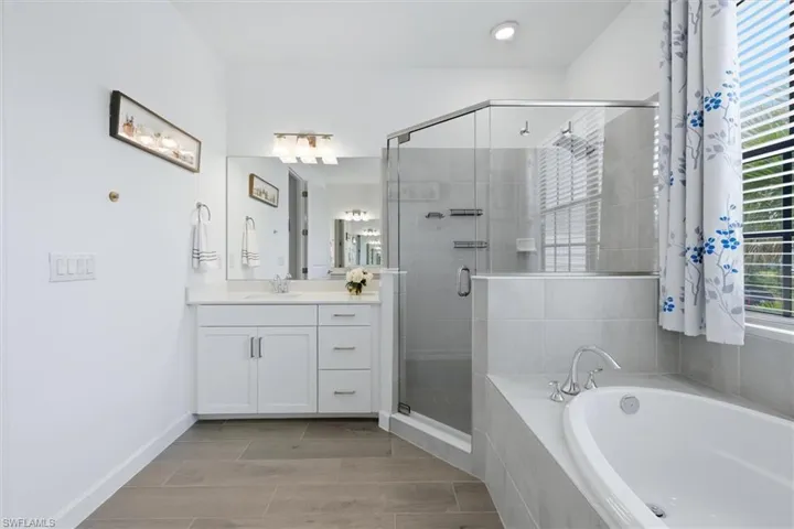 Bathroom featuring a white vanity with a single sink, an enclosed glass shower, a built-in soaking tub, wood-finish flooring, and a large window with privacy blinds