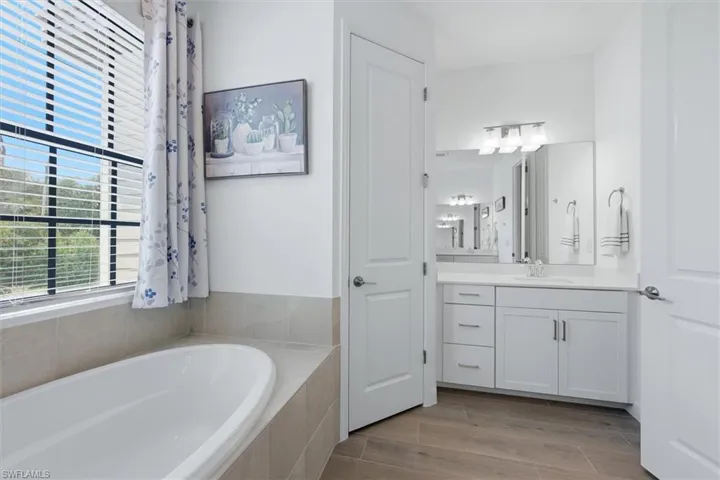 Soaking tub with tiled surround, large window with blinds, dual sink vanity with white cabinetry, wall-mounted mirror, and wood-finish flooring