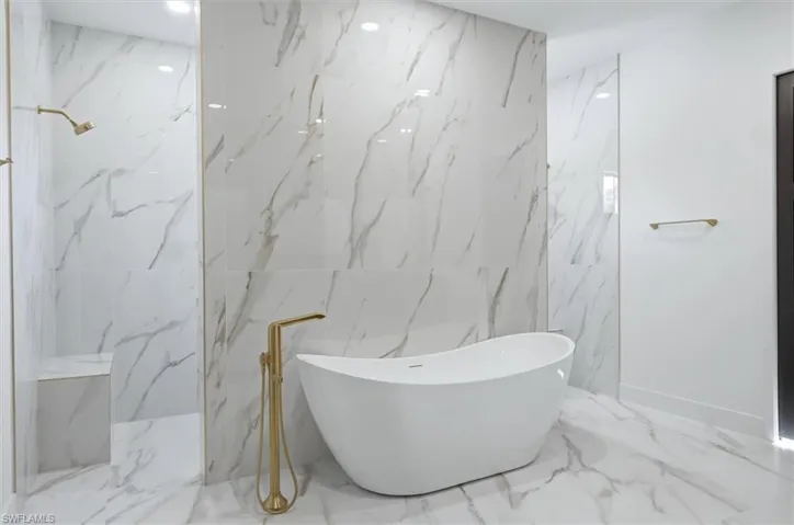 Freestanding soaking tub with a floor-mounted gold-tone faucet, walk-in shower featuring a built-in bench, and large format marble-look tile flooring and walls