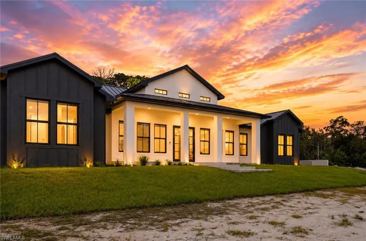 Modern farmhouse residence featuring a striking black and cream exterior with board-and-batten siding, expansive front-facing windows with illuminated frames, gabled roofline with clerestory detailing, manicured lawn, and mature landscape framing on a generous lot