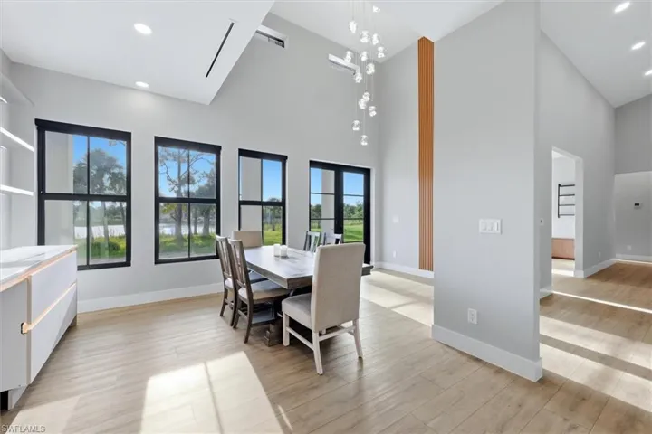 Modern open-concept dining and kitchen area featuring floor-to-ceiling black-framed windows with expansive natural light, wood-finish flooring throughout, a contemporary pendant light fixture, a vertical wood-grain accent column, and seamless sightlines to additional living spaces
