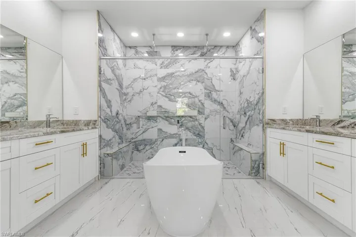 Bathroom featuring two vanities, a marble finish shower, a freestanding bath, recessed lighting, and light marble finish floors