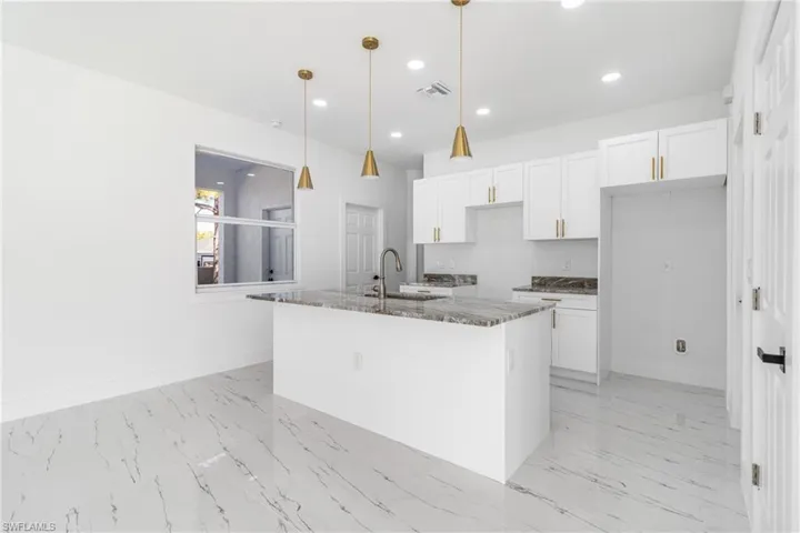 Kitchen featuring white cabinetry, dark stone countertops, hanging light fixtures, a kitchen island with sink, and light marble finish flooring