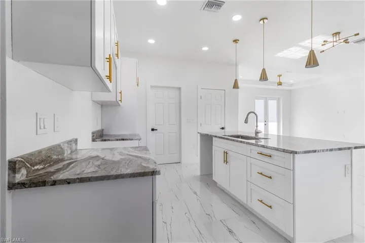 Kitchen featuring white cabinets, dark stone counters, light marble finish floors, an island with sink, and decorative light fixtures